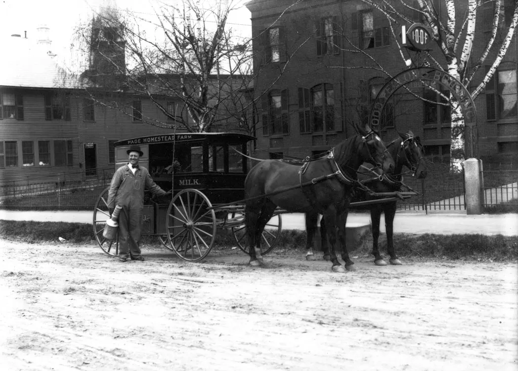 A milkman in overalls holding a milk can stands beside a two-horse wagon marked “Page Homestead Farm” and “Milk” on a dirt street