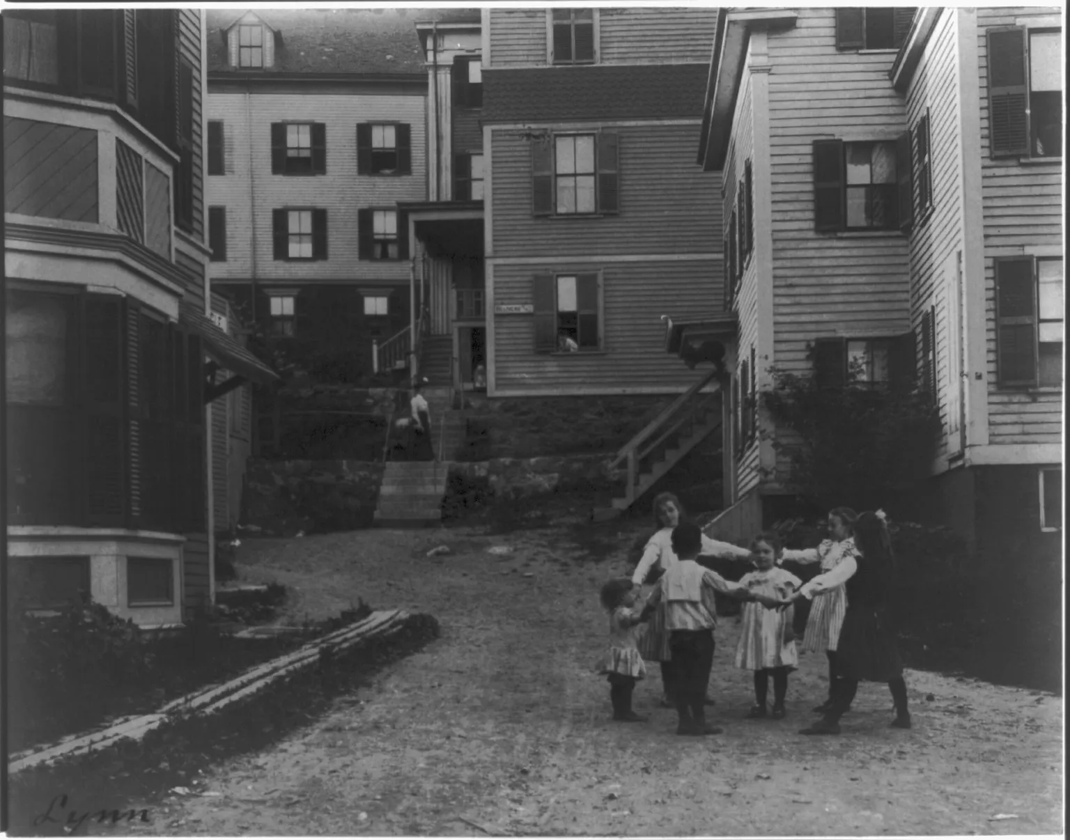 Six small children hold hands in a ring on a dirt lane between frame houses